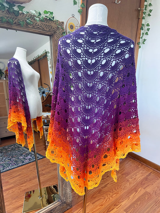 A crocheted shawl displayed on a white mannequin in an indoor room. The garment has an ombré color gradient transitioning from purple at the top to orange and yellow at the bottom, with an intricate openwork lace pattern and scalloped edges.