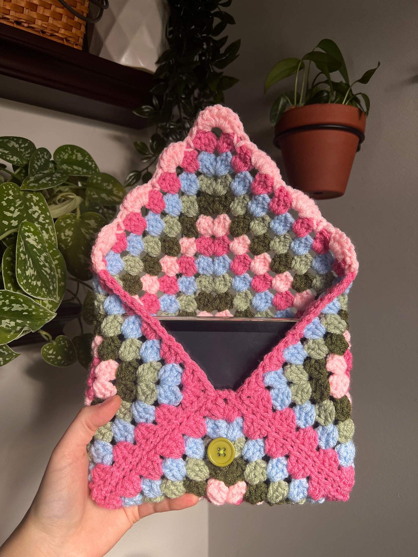 Colorful crocheted book sleeve held by a hand with plants in the background