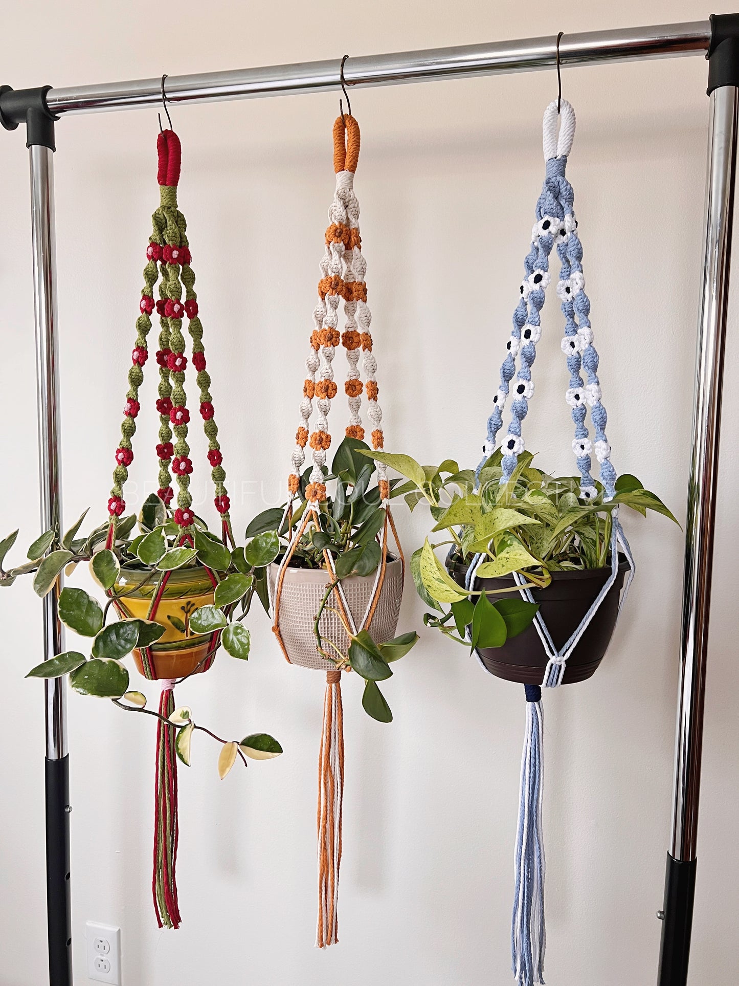 Three macrame plant hangers with colorful flowers on a garment rack against a white wall.