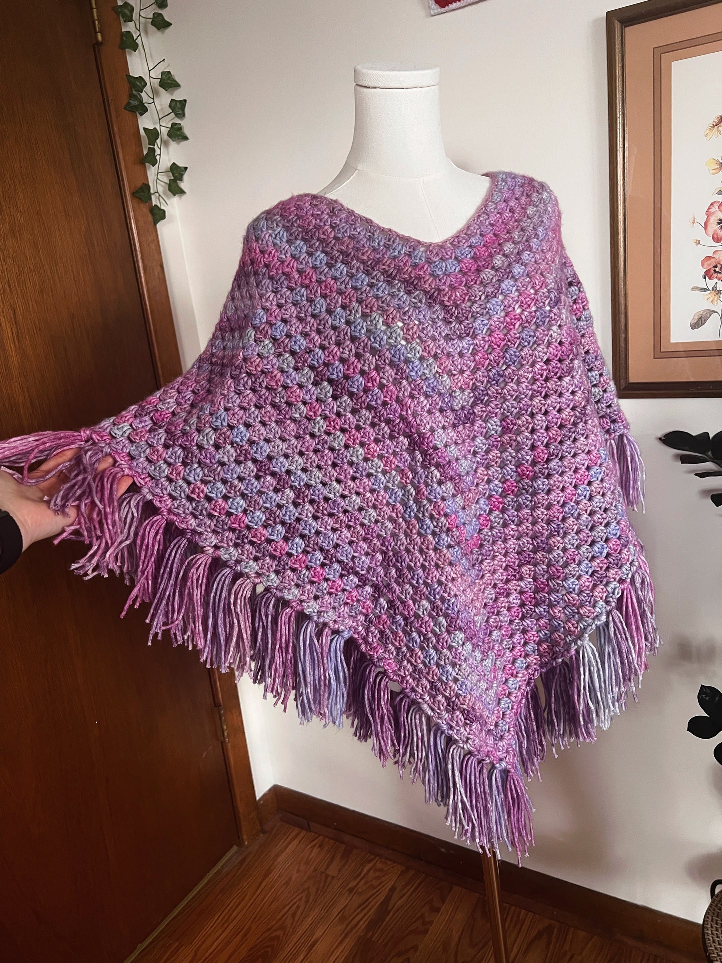 Purple crocheted poncho on a mannequin against a wall with a framed picture.