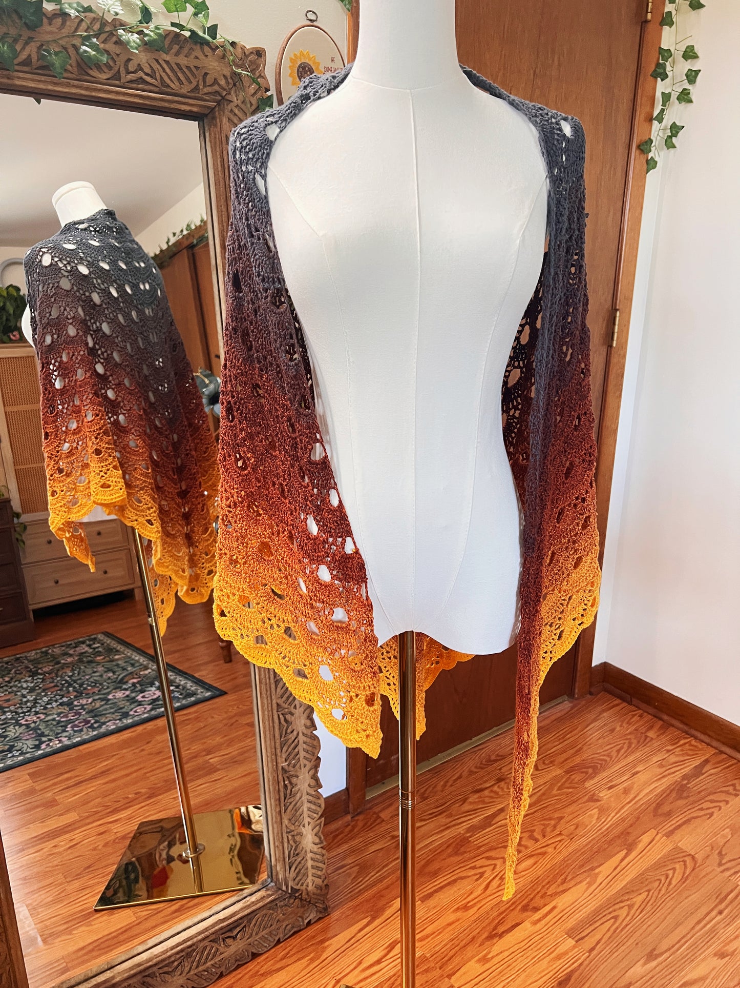 A hand-crocheted lace shawl with an ombre gradient from dark gray to orange and yellow, displayed on a white mannequin torso in a home setting with a wooden floor and mirror.