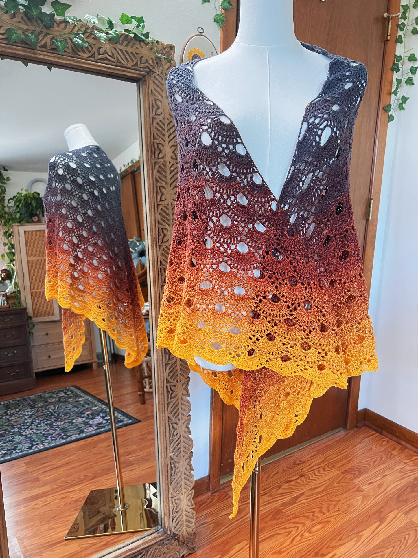 A crocheted shawl with a gradient color transition from grey to orange and bright yellow, displayed on a white mannequin in front of a mirror showing its reflection.