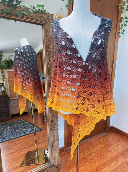 A crocheted shawl with a gradient color transition from grey to orange and bright yellow, displayed on a white mannequin in front of a mirror showing its reflection.