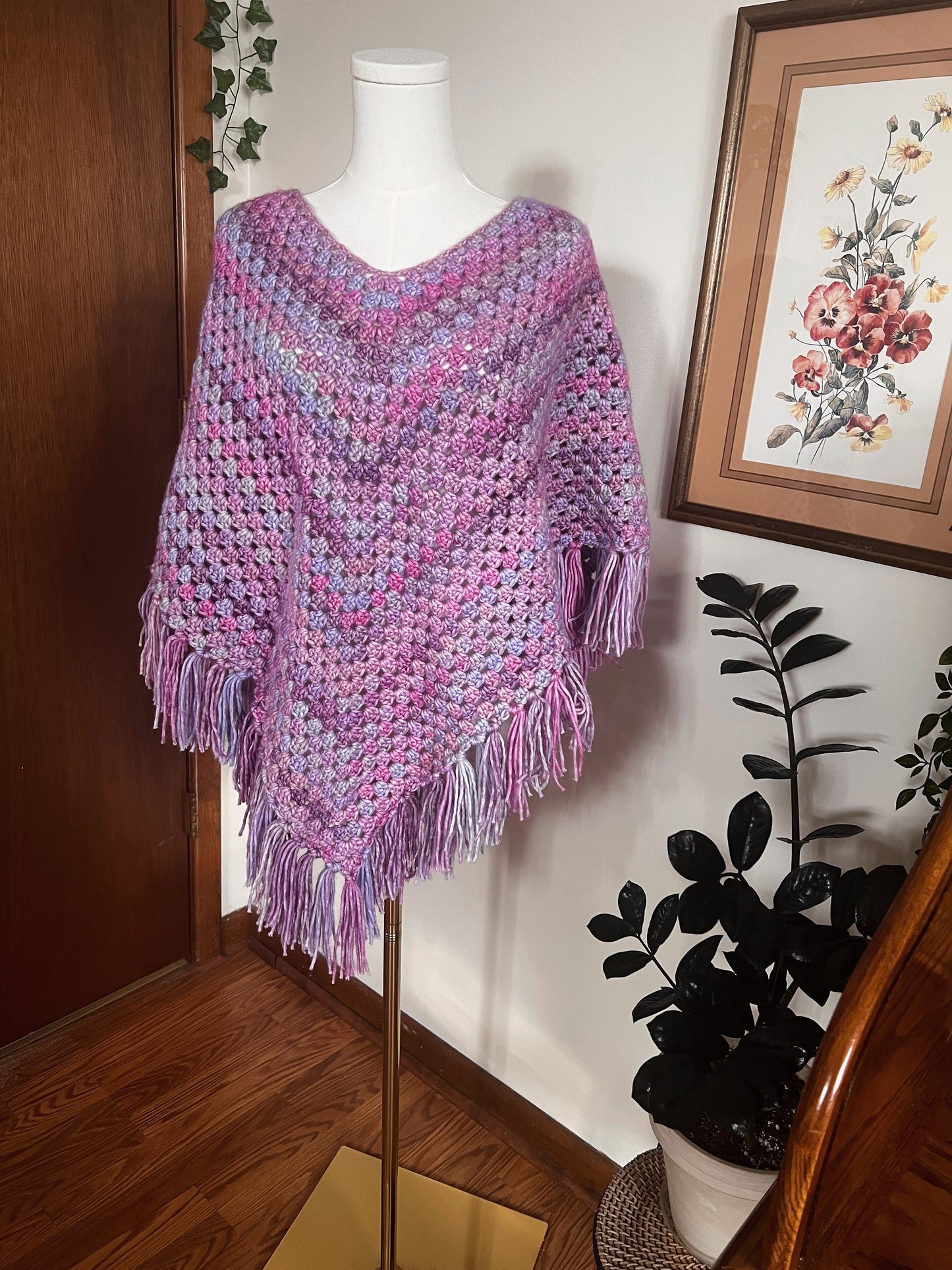 Purple crochet poncho on a mannequin against a wall with a framed picture and plant.