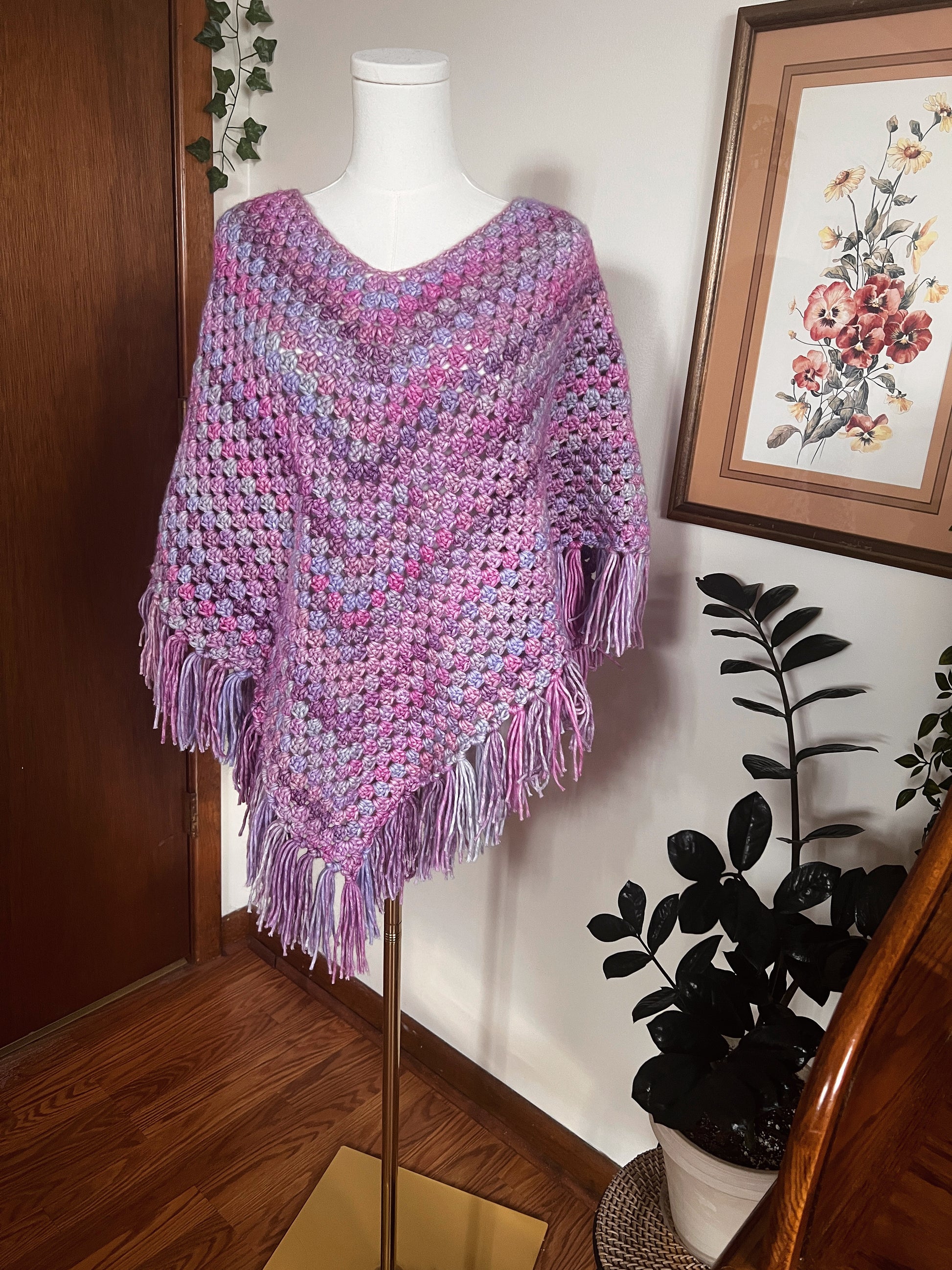 Purple crochet poncho on a mannequin against a wall with a framed picture and plant.