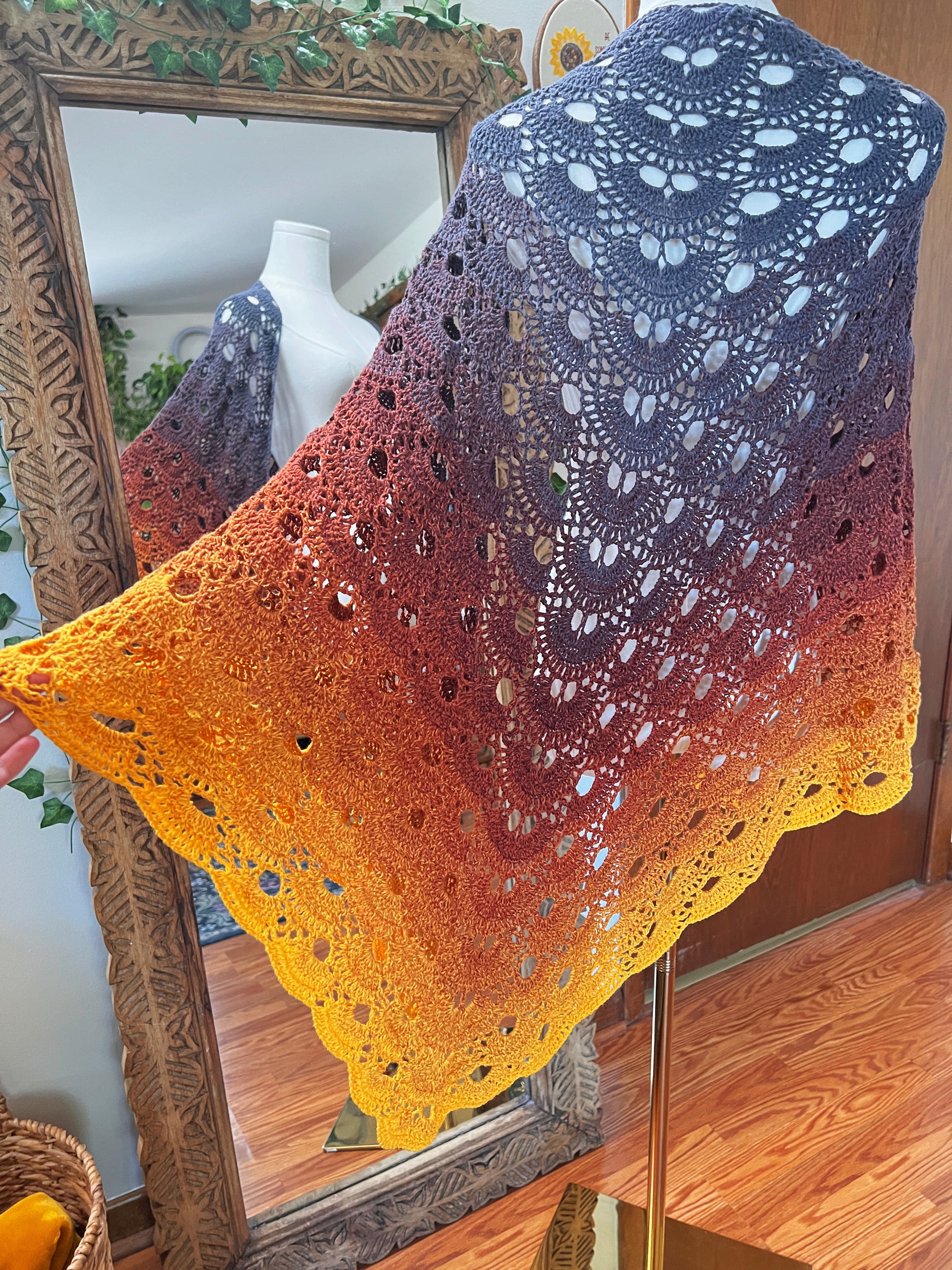 A large open-crochet lace shawl with a warm gradient color transition from grey at the top to brown and bright yellow at the bottom, draped over a mannequin in front of a large ornate wooden mirror.
