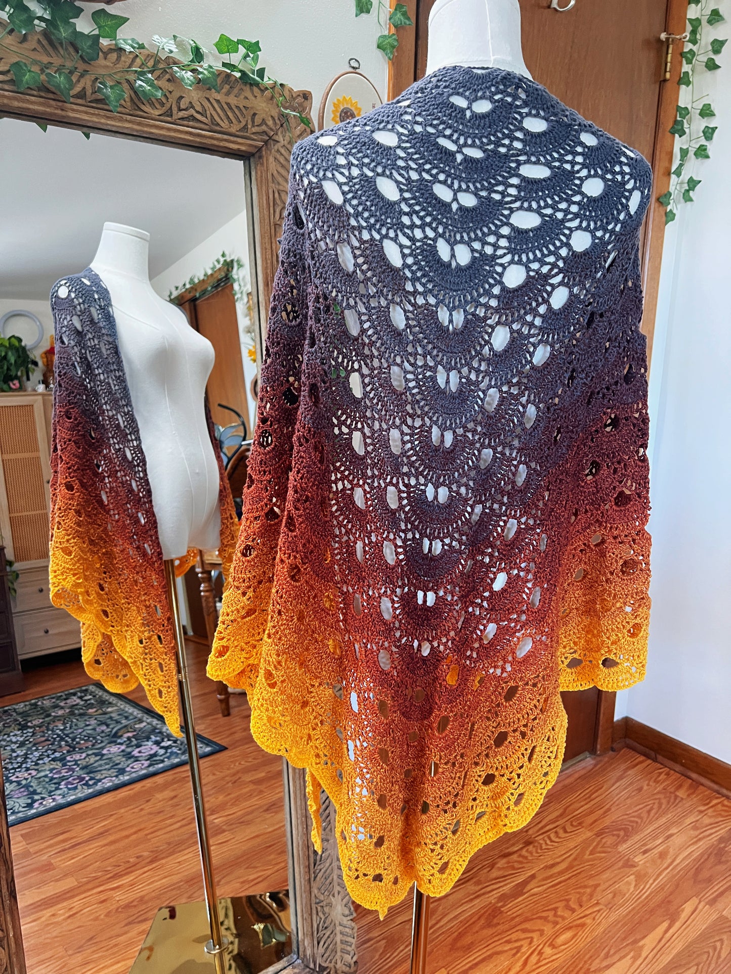 A hand-crocheted shawl with a gradient color transition from dark charcoal gray to orange and bright yellow, displayed on a mannequin in an indoor room with a mirror reflecting its side view.