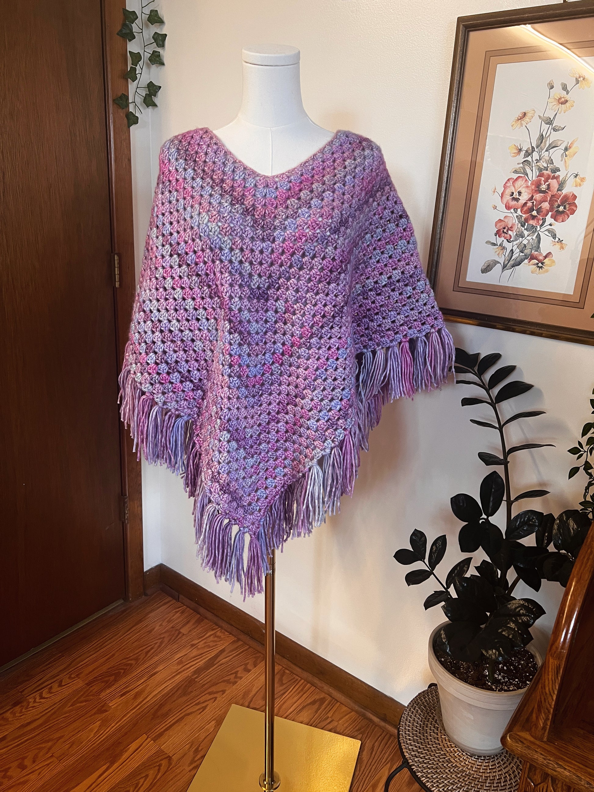 Purple crocheted poncho on a mannequin against a wall with a plant and framed picture.