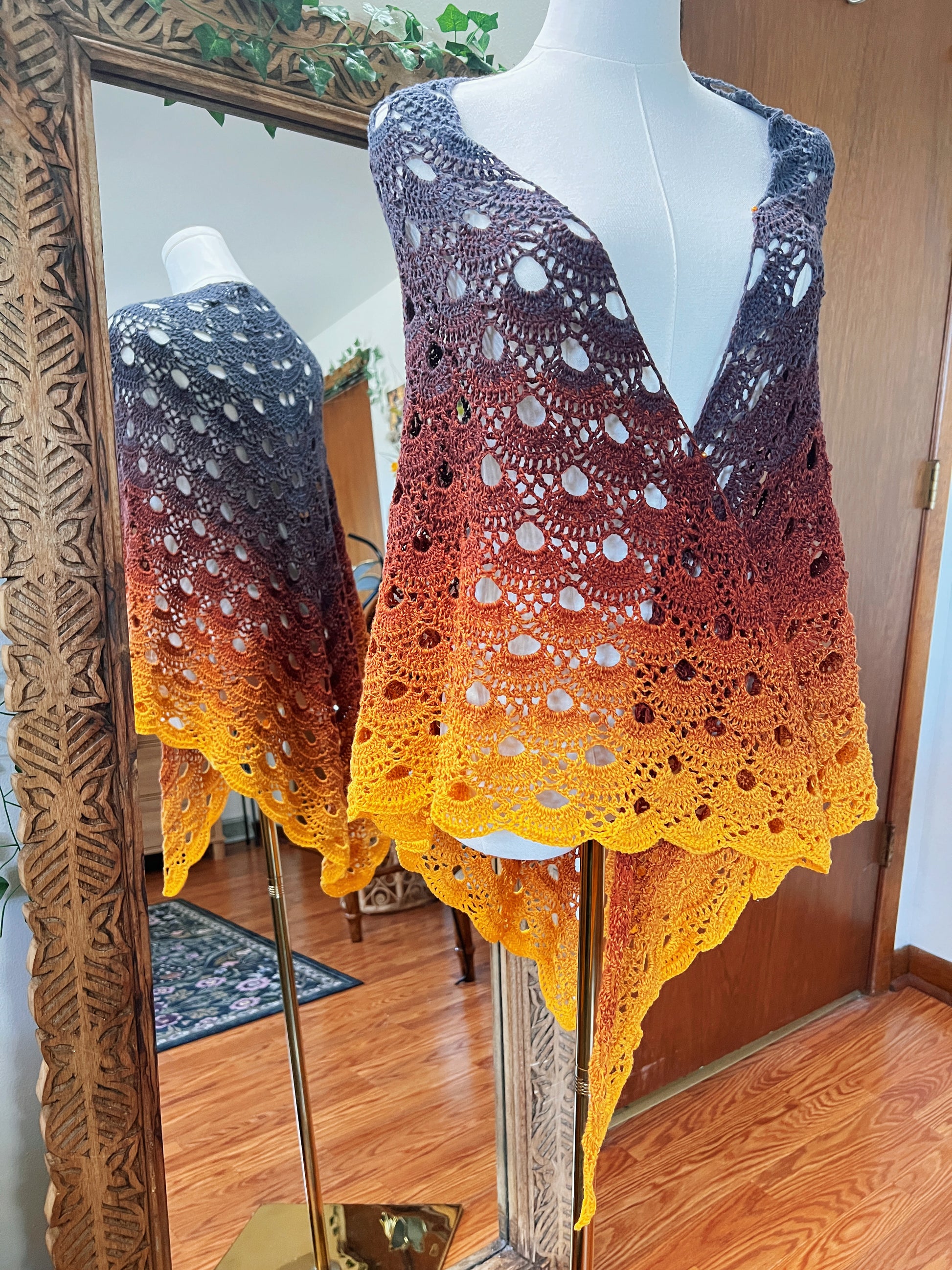A crocheted openwork shawl draped on a white mannequin torso, shown indoors with a mirror reflecting the back side. The color gradient transitions from dark charcoal at the top through rust red and orange to bright yellow at the hem.