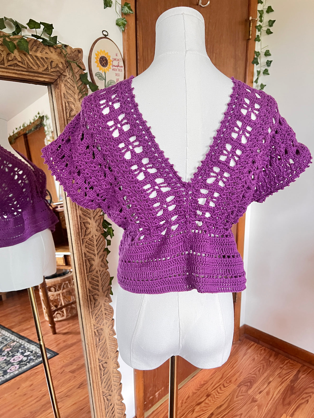 A purple crocheted short-sleeve top displayed on a white dress form mannequin, with a mirror in the background reflecting the garment and showing a cozy indoor room setting.