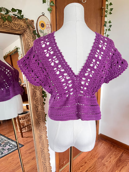 A purple crocheted short-sleeve top displayed on a white dress form mannequin, with a mirror in the background reflecting the garment and showing a cozy indoor room setting.