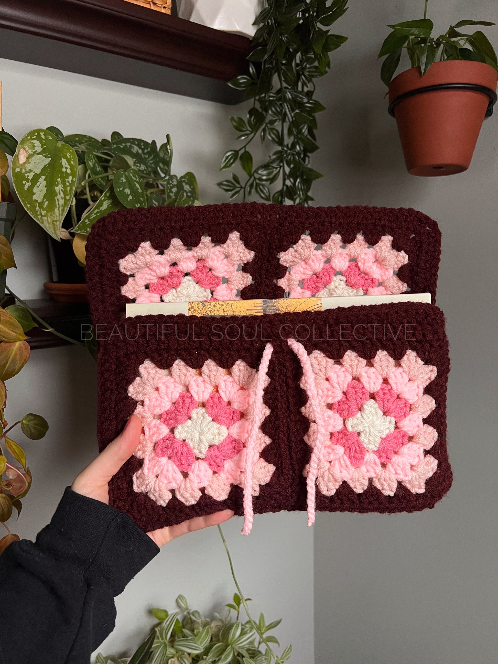 Handmade crochet book sleeve with pink and white patterns held by a person, with plants in the background.