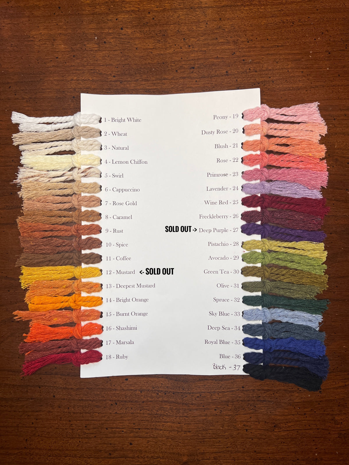 A printed color chart sheet showing 28 macrame cord swatches arranged in two columns on a dark wooden table, with some swatches physically attached to the page.