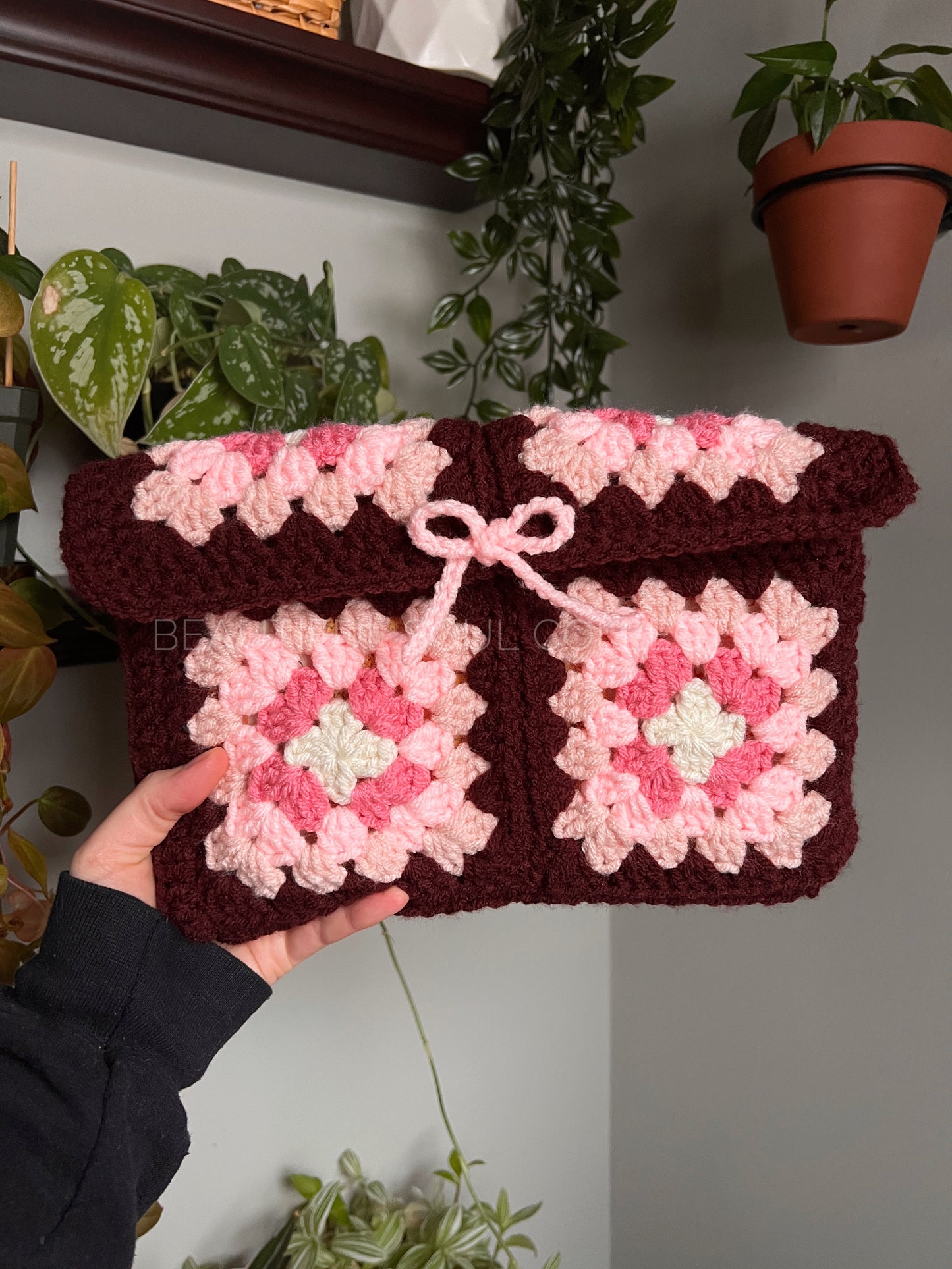 Handmade crochet book sleeve with pink and white pattern held by a person, with plants in the background.