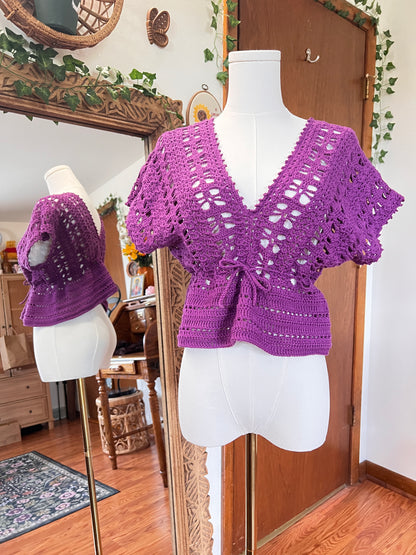 A purple crocheted short-sleeve top displayed on a white dress form mannequin, with a mirror in the background reflecting the garment and showing a cozy indoor room setting.