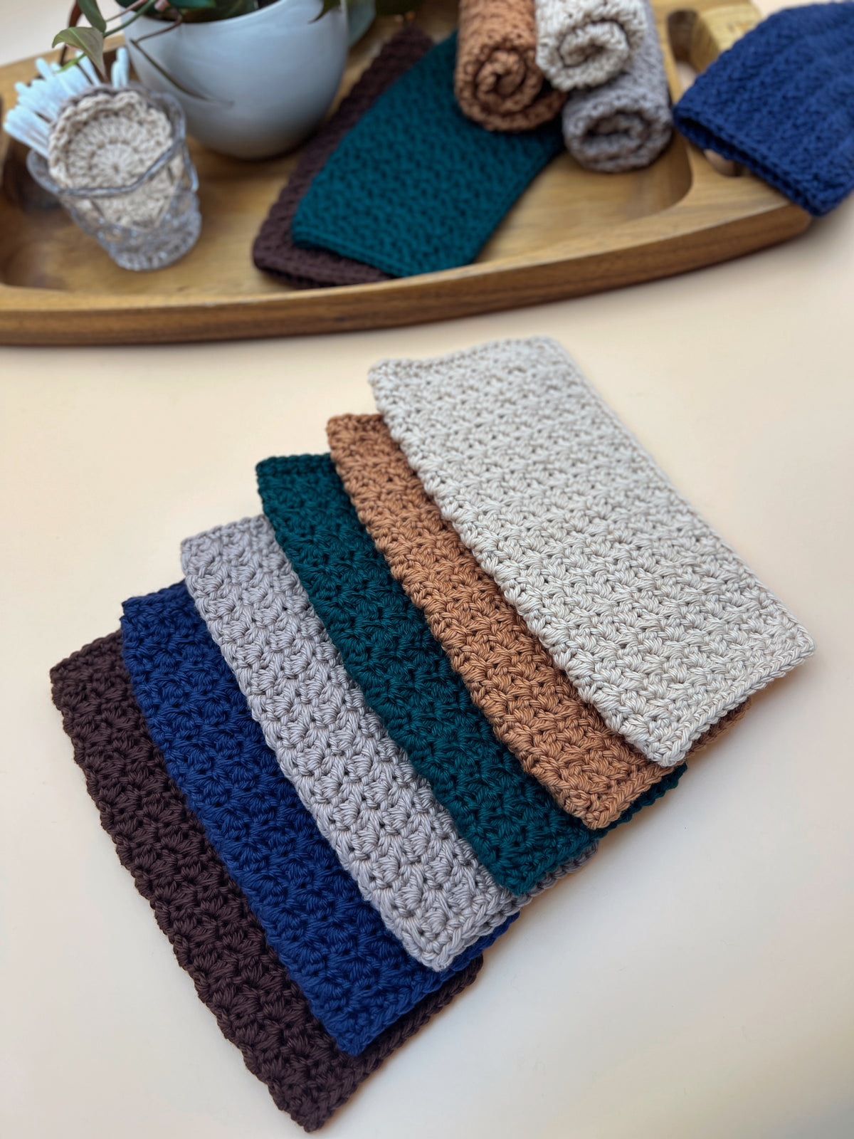 A set of six textured crochet washcloths in different colors (dark brown, navy, light gray, teal, terracotta, and off-white) arranged in a fan shape on a light beige surface. In the background, a wooden tray holds more folded fabric swatches and a small glass jar.