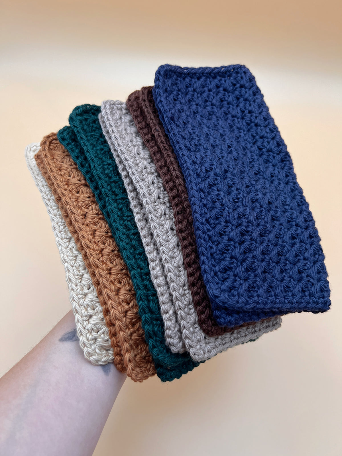 Six crochet washcloths held by a person in tan, orange, teal, taupe, brown and navy blue