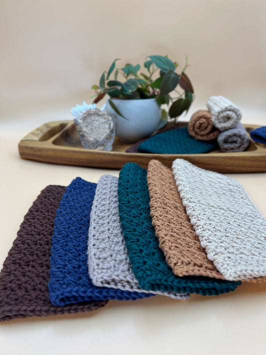 Six textured crocheted dishcloths in brown, blue, light gray, teal, tan, and off-white, arranged in a fan shape in the foreground. A wooden tray with a small potted plant and rolled towels is visible in the softly blurred background.
