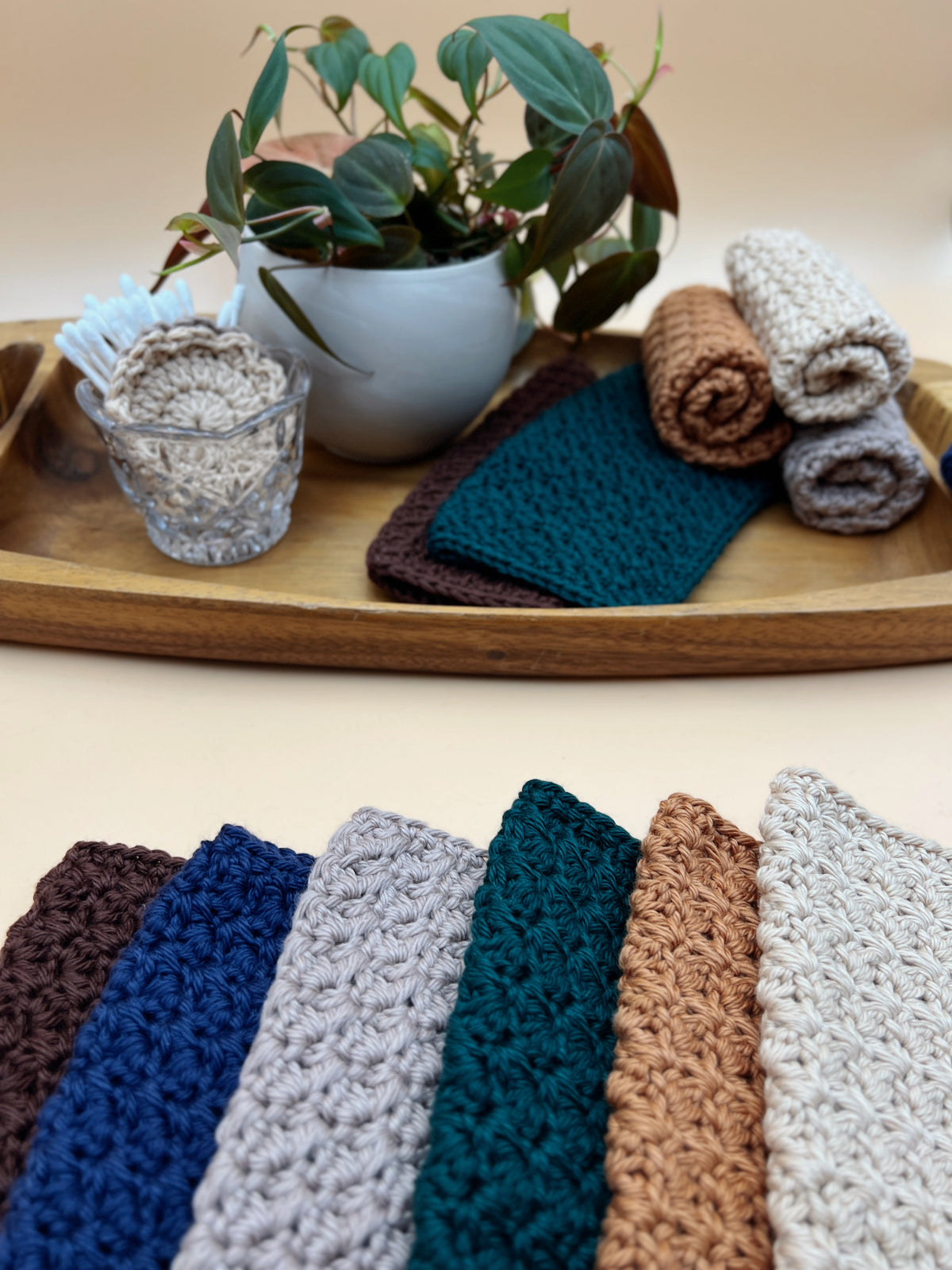 A set of textured crochet washcloths in multiple colors, shown both folded on a wooden tray and arranged in a fan at the bottom. The visible colors include dark brown, navy, light gray, teal, tan, and off-white.