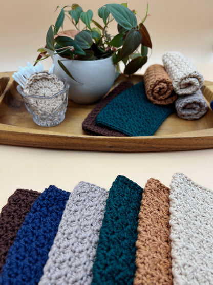 A set of textured crochet washcloths in multiple colors, shown both folded on a wooden tray and arranged in a fan at the bottom. The visible colors include dark brown, navy, light gray, teal, tan, and off-white.