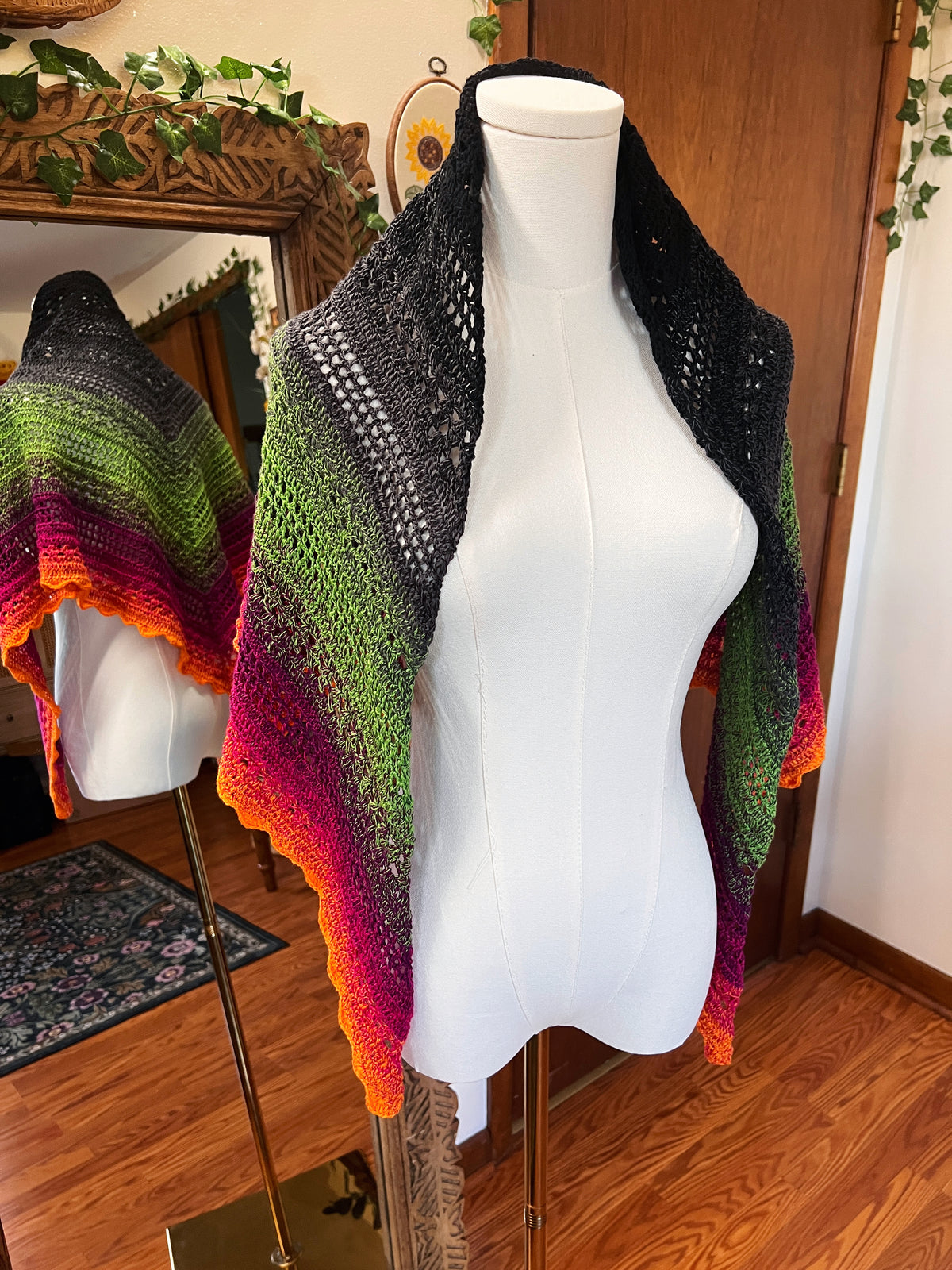 Crochet shawl in a gradient of black, grey, green, pink and orange on a mannequin in front of a mirror