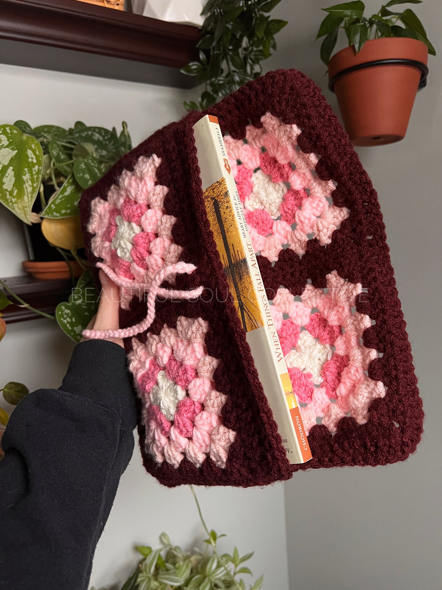 Person holding a crocheted e-reader sleeve with pink and white patterns against a home interior background.