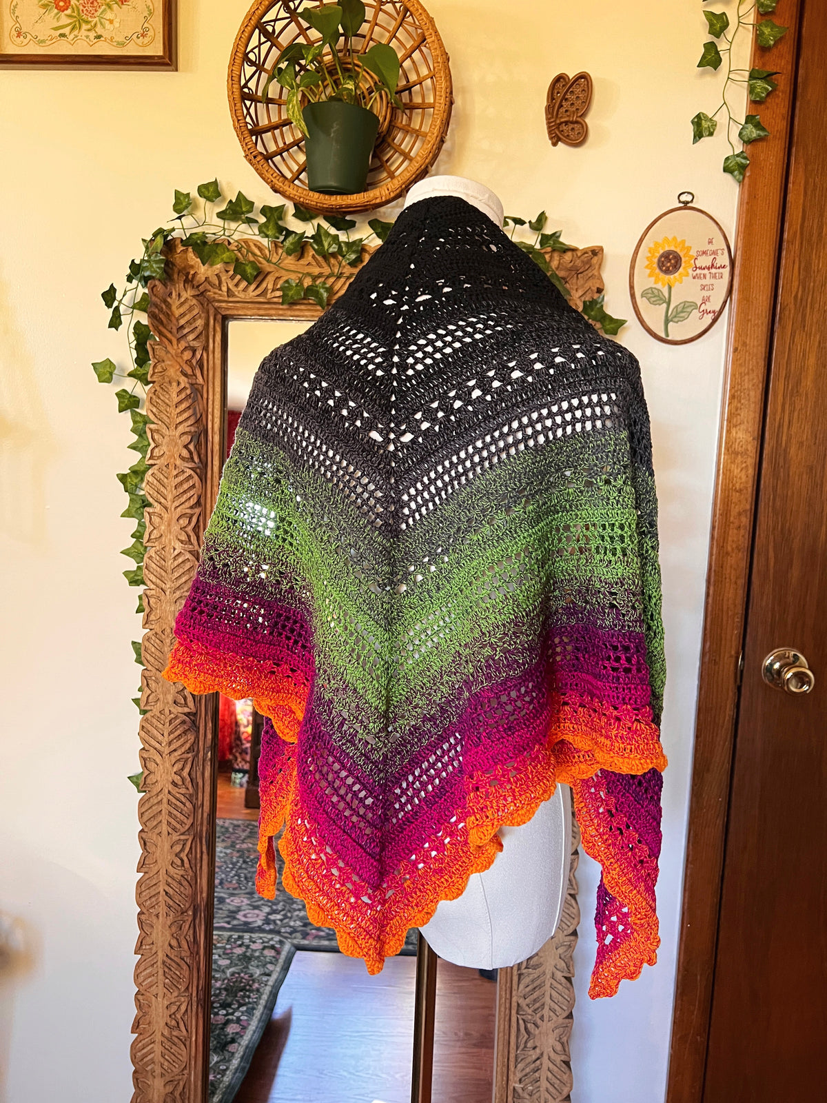 Crochet shawl in a gradient of black, grey, green, pink and orange on a mannequin in front of a mirror
