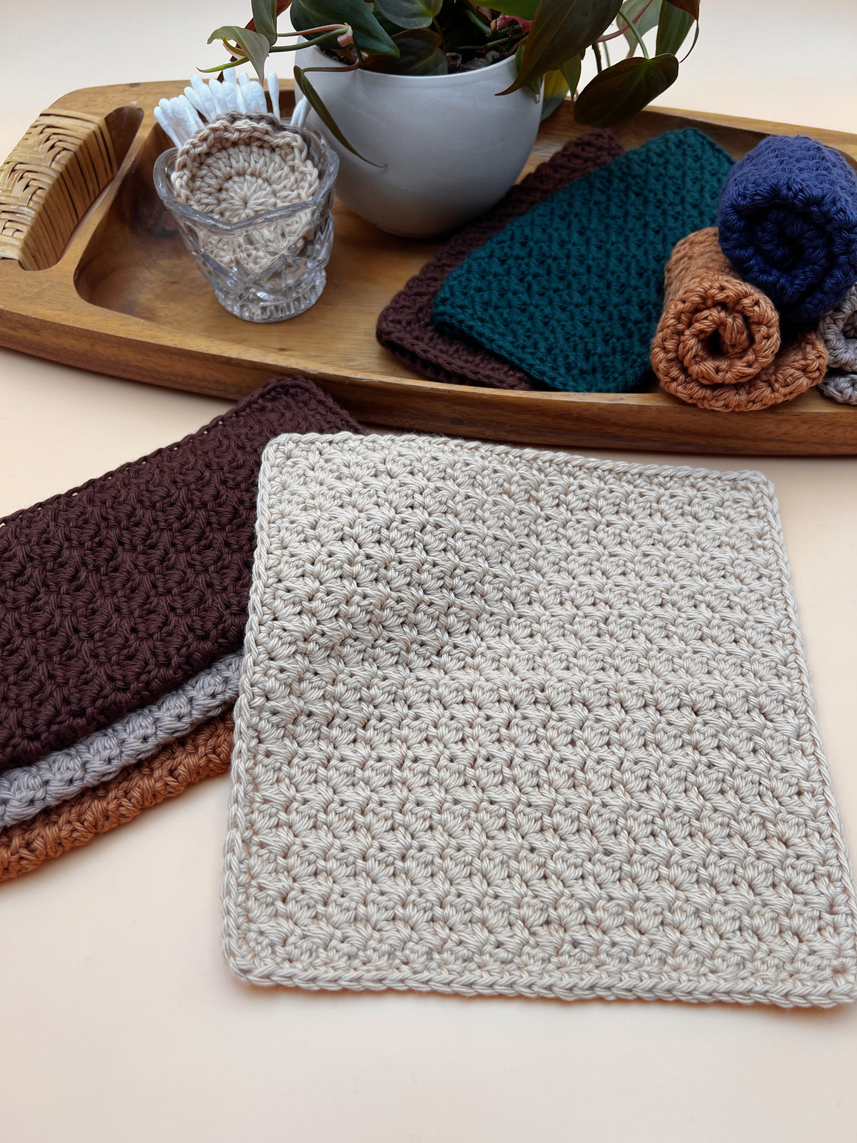 A square crocheted dishcloth in light beige is displayed in the foreground, with other folded and rolled crocheted dishcloths in dark brown, gray, teal, and brown arranged on a wooden tray and beige surface.