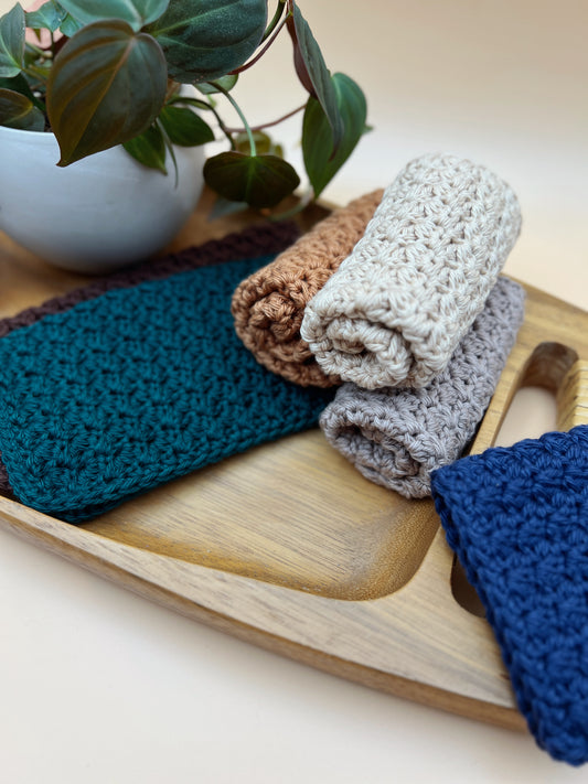 Four textured crochet washcloths in teal, brown, light gray, and navy, folded and stacked on a wooden tray next to a potted plant.