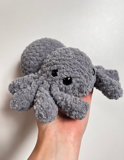 Gray plush toy resembling a spider held in a hand against a plain background
