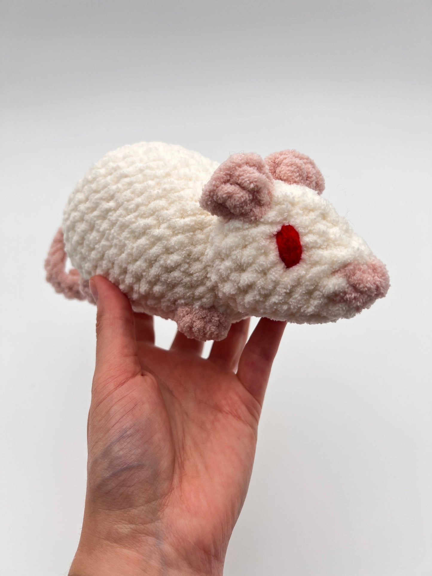 Hand holding a small white crocheted rat toy with red eyes against a plain background