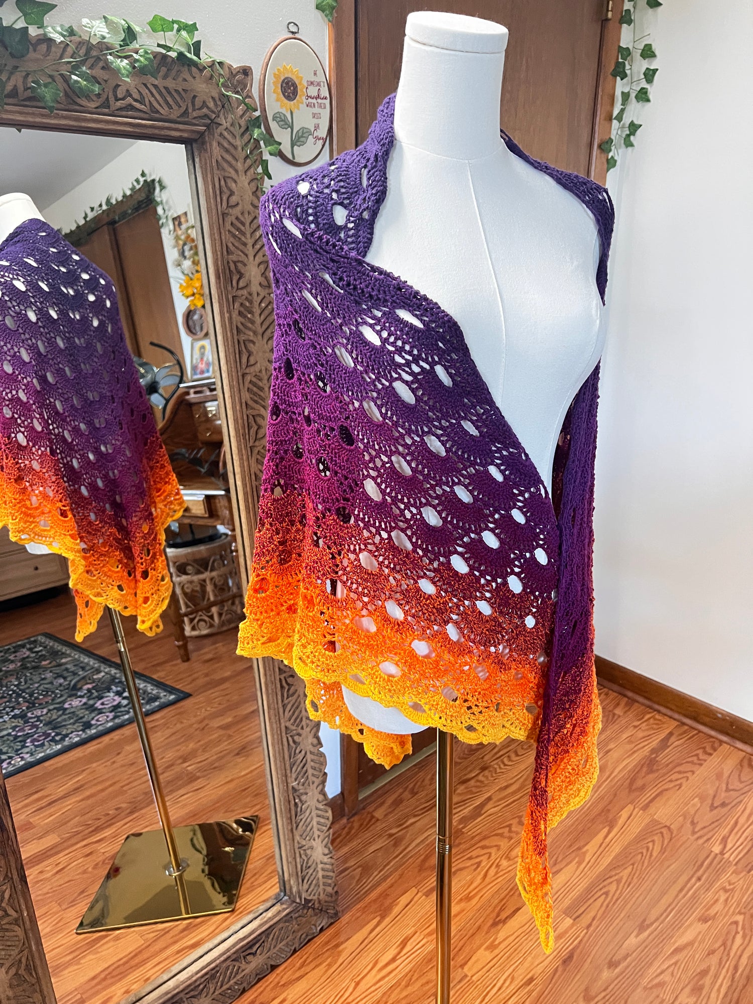 A crocheted shawl with an orange and purple gradient, displayed on a white mannequin torso in an indoor room with wooden floors and a mirror.