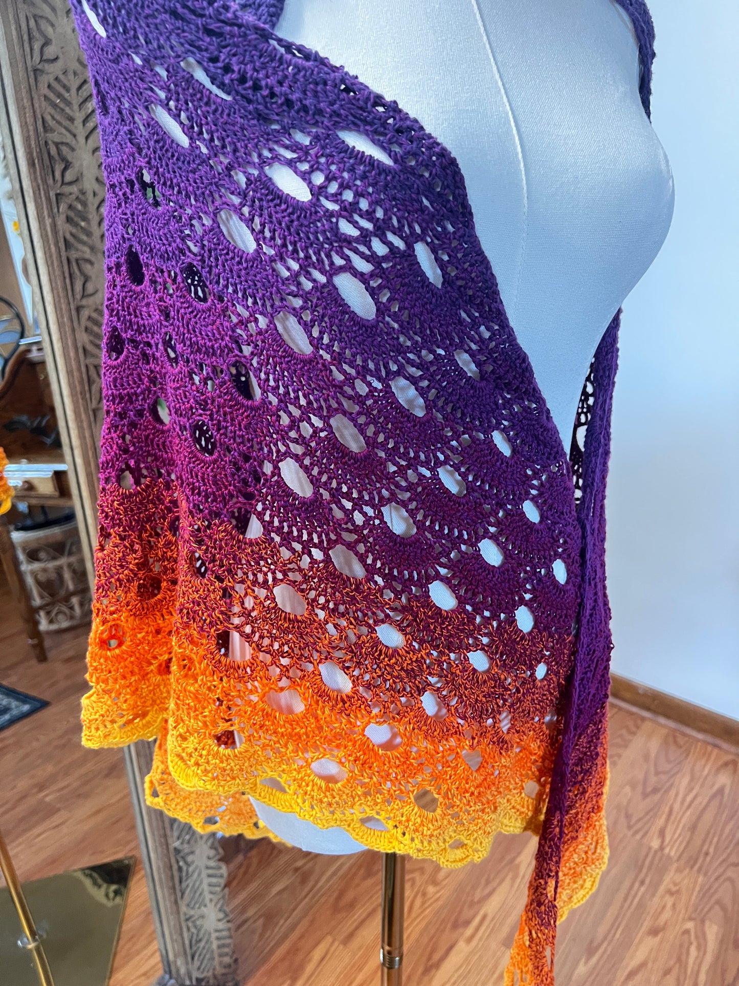 A hand-crocheted openwork shawl with a gradient color transition from purple to orange to yellow, displayed on a white mannequin torso in an indoor setting.
