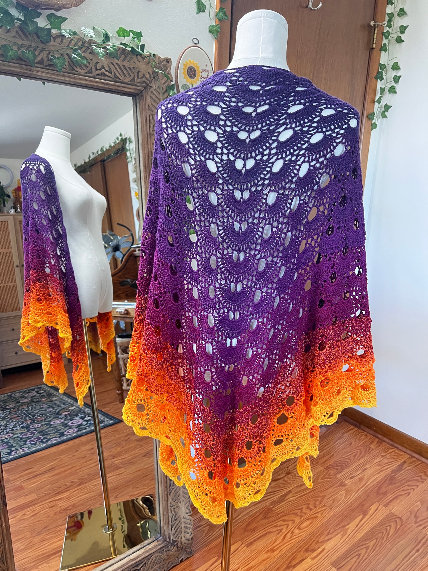 A crocheted shawl displayed on a white mannequin in an indoor room. The garment has an ombré color gradient transitioning from purple at the top to orange and yellow at the bottom, with an intricate openwork lace pattern and scalloped edges.