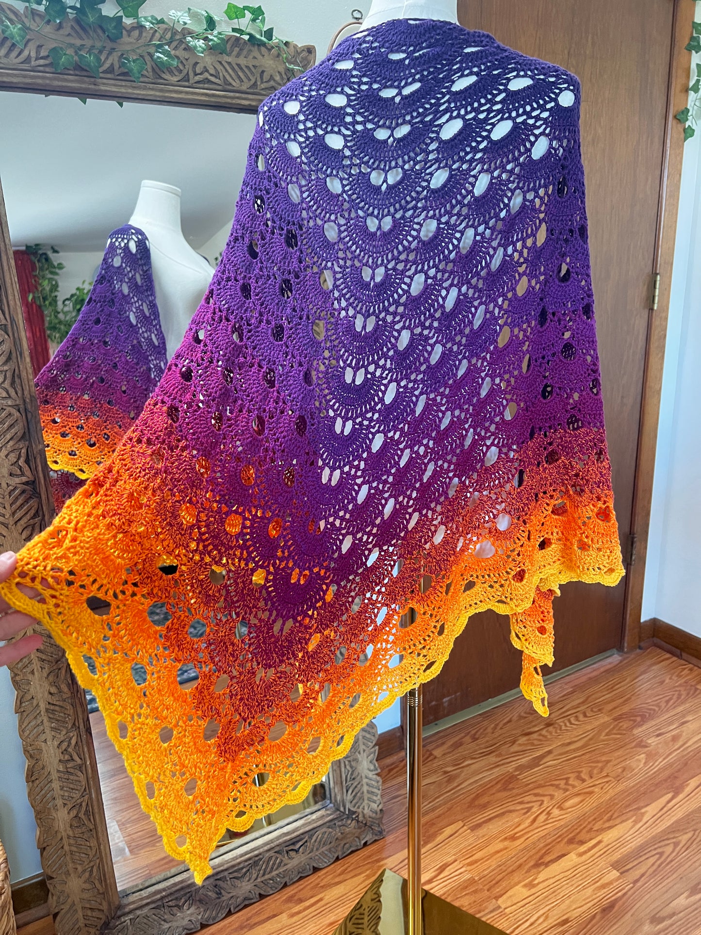 A handmade crochet shawl with a purple-to-yellow gradient, draped over a mannequin in an indoor room with wooden floors and a mirror.