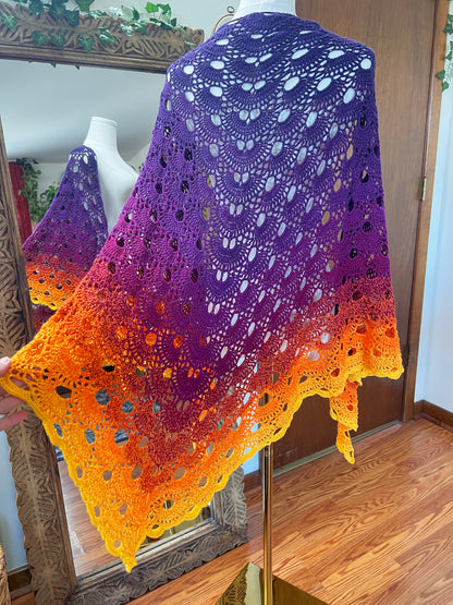 A handmade crochet shawl with a purple-to-yellow gradient, draped over a mannequin in an indoor room with wooden floors and a mirror.