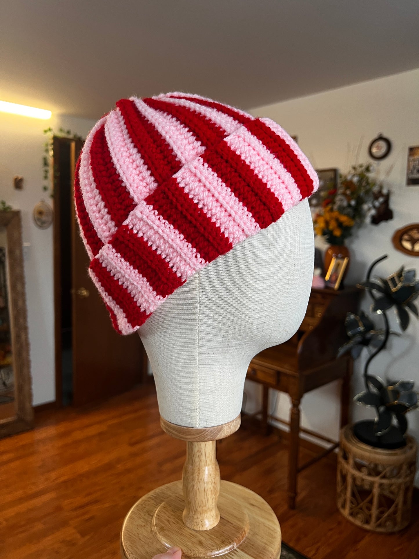 Red and pink striped crochet hat on a mannequin head against a home interior background.