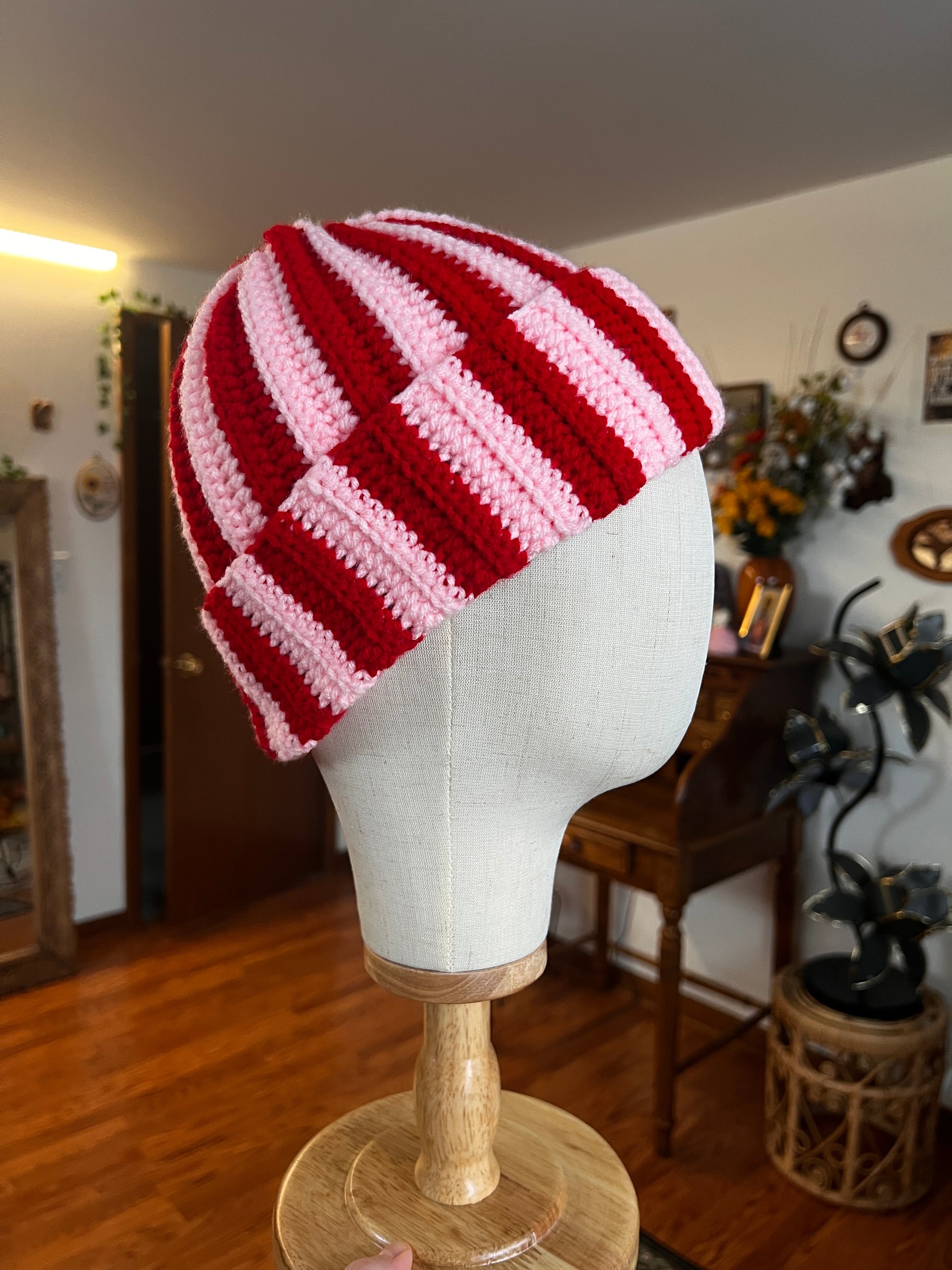Red and pink striped crochet hat on a mannequin head against a home interior background.
