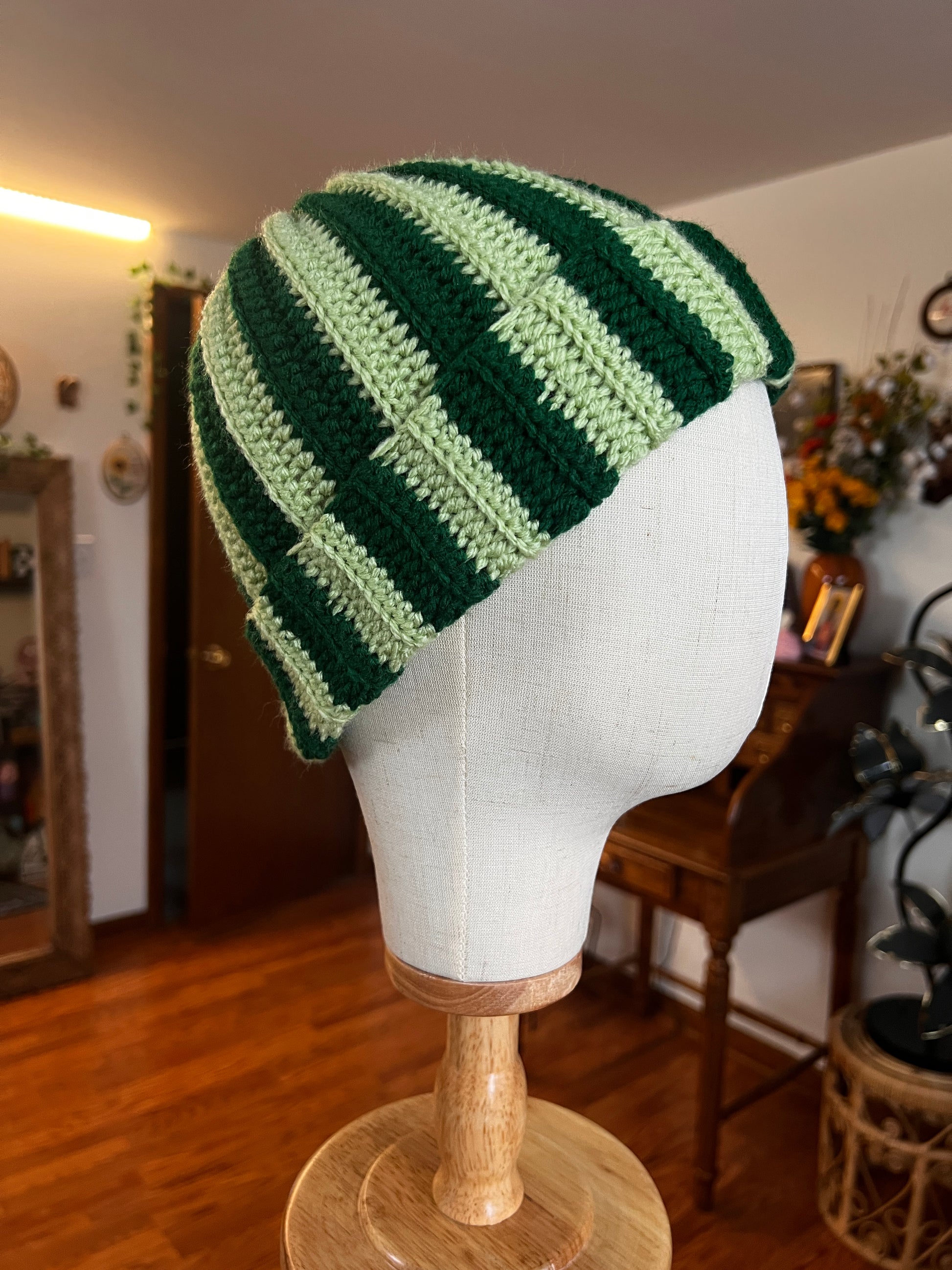 Green striped crocheted hat on a mannequin head against a blurred indoor background.