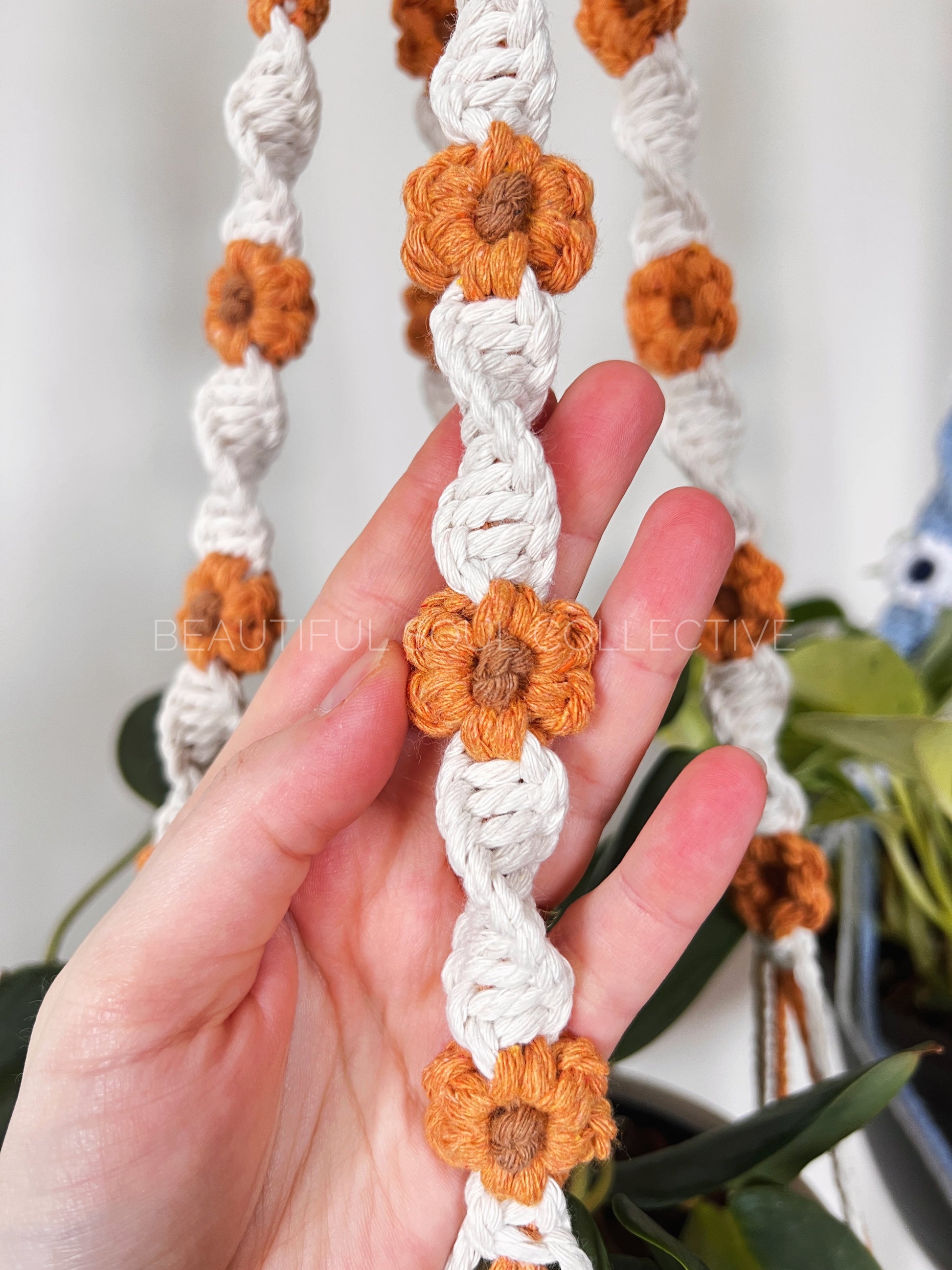 Hand holding a macrame plant hanger with orange flowers and white strings.