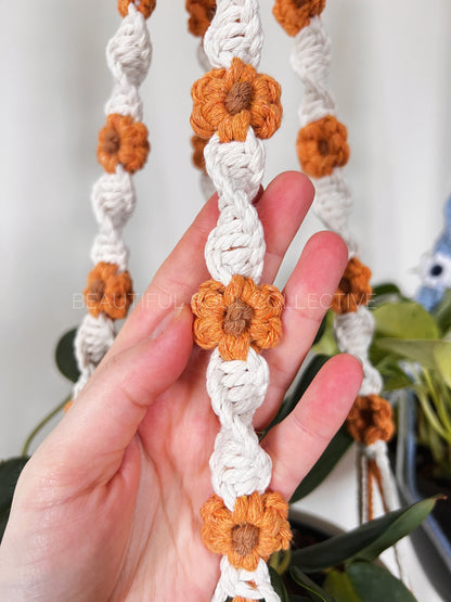 Hand holding a macrame plant hanger with orange flowers and white strings.