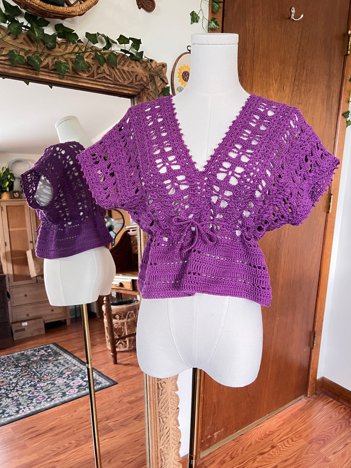 A purple crocheted short-sleeve top displayed on a white dress form mannequin, with a mirror in the background reflecting the garment and showing a cozy indoor room setting.