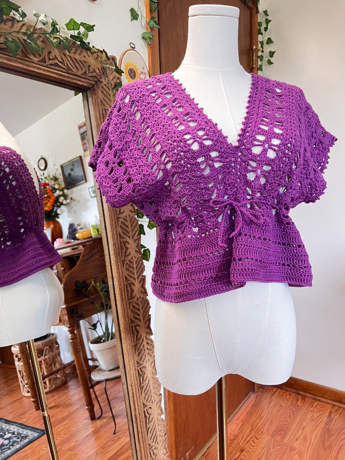 A purple crocheted short-sleeve top displayed on a white dress form mannequin, with a mirror in the background reflecting the garment and showing a cozy indoor room setting.