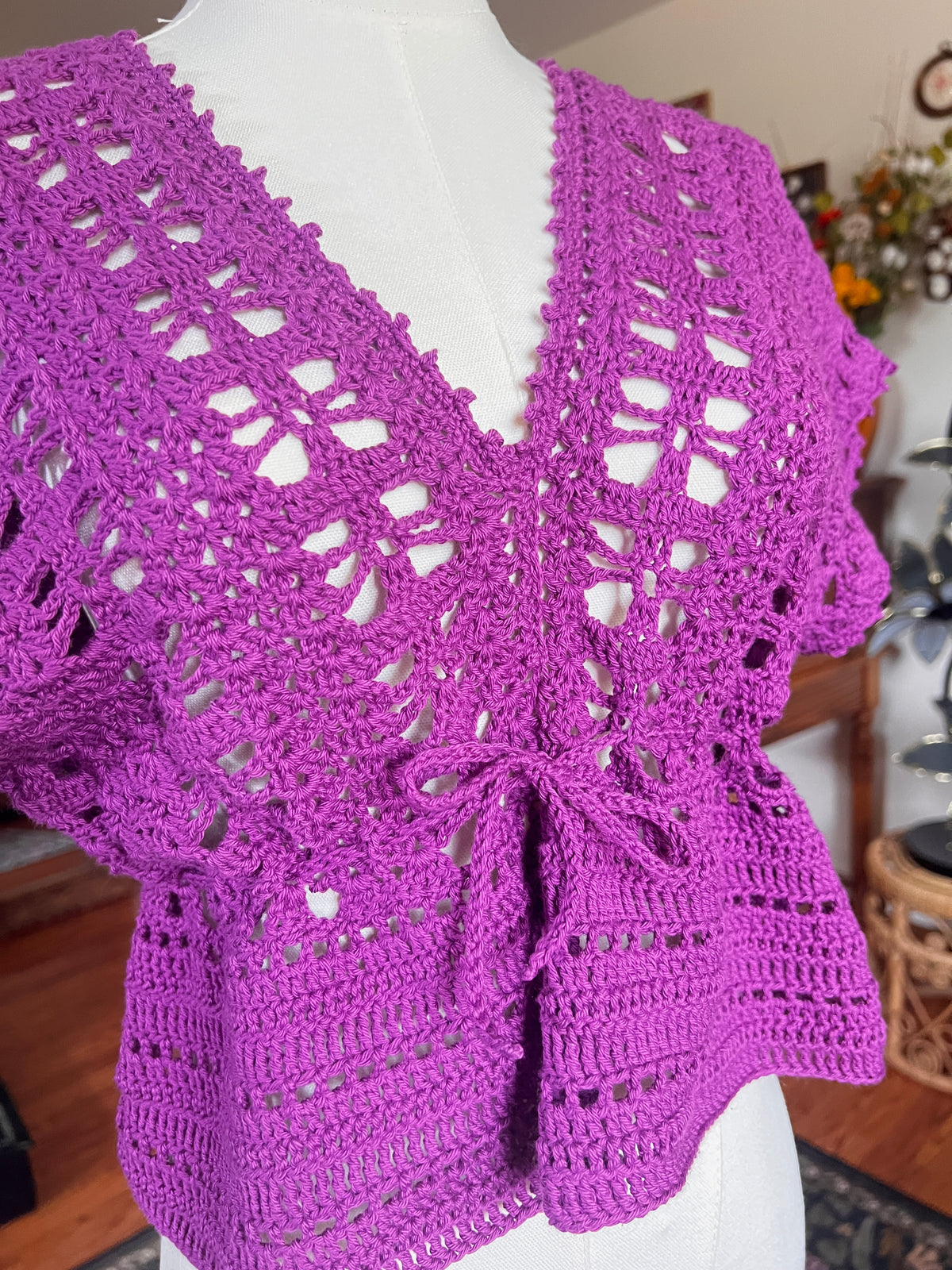 A bright purple hand-crocheted top with a V-neck and tie-front detail, displayed on a white mannequin torso in an indoor setting.