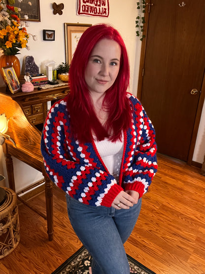 Person wearing a red, white, and blue crochet sweater in a room with wooden floors and furniture.