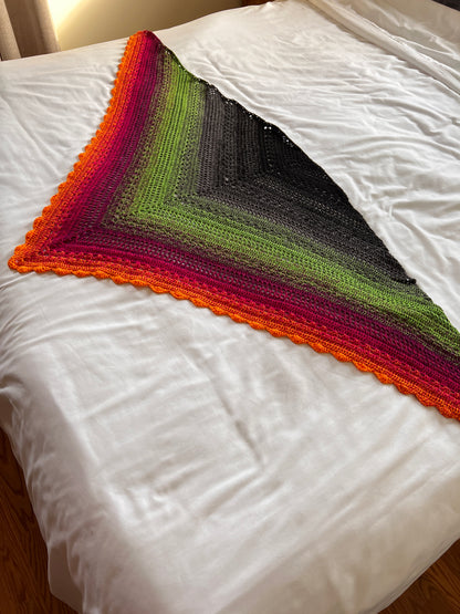 Crochet triangle shawl in a gradient of black, grey, green, pink and orange laid flat on a white bed